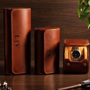 Puede incluir: Tres estuches cilíndricos para relojes de cuero marrón con acabado liso. Dos estuches están cerrados y presentan monogramas verticales, uno con L|V y otro con T.S. El tercer estuche está abierto, revelando un reloj de pulsera plateado con esfera negra.