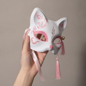 May include: A white fox mask featuring pink painted swirl patterns around the eye openings and ears. The mask includes a small gold bell attached to the right side, accompanied by a light pink fabric tassel and a matching pink satin ribbon tie.
