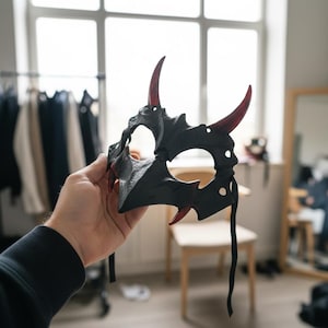 May include: A black mask featuring a pointed, angular design with two prominent upward-curving horns and smaller downward-pointing spikes. The mask includes translucent red accents on the horns and lower spikes, with black fabric ties attached.