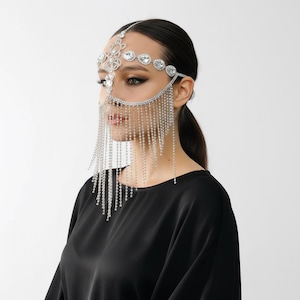 May include: A silver-toned face accessory featuring a decorative headpiece made of clear, faceted crystals arranged in geometric patterns across the forehead and bridge of the nose, with long, dangling crystal fringe covering the lower face.