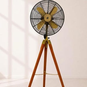 May include: A floor fan featuring a circular black wire cage and four gold-toned blades mounted on a tall, three-legged wooden tripod stand. The central hub displays a circular emblem with a ship graphic and text, connected by a gold chain.
