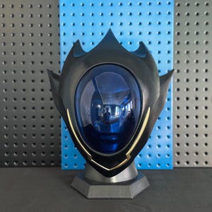 Puede incluir: Un soporte para casco en negro mate con una visera central ovalada de color azul profundo translúcido. El casco tiene una parte superior puntiaguda en forma de corona y protuberancias laterales angulares, con finos bordes dorados en la visera.