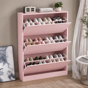 Puede incluir: Un zapatero alto de color rosa claro con tres compartimentos de almacenamiento abatibles, cada uno con varios pares de zapatos. La superficie superior sostiene un reloj digital que marca las 09:09, dos libros apilados y una planta pequeña.
