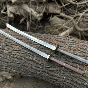 Op de afbeelding: Twee zwaarden met rechte, gemêleerde metalen bladen, goudkleurige kragen en donkere handvatten. Het ene zwaard heeft een donkerbruin houten handvat, terwijl het andere een zwart getextureerd handvat heeft. Beide handvatten zijn met metalen pinnen.