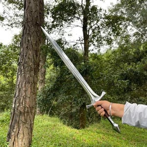 Peut inclure: Une épée en métal argenté à double tranchant avec une pointe acérée, une garde transversale et un manche marron foncé. La lame présente une arête centrale sur toute sa longueur et la poignée se termine par un pommeau pointu, tenue par une main.