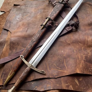 May include: A double-edged steel sword with a polished blade, a brass crossguard, and a wooden hilt featuring a brass pommel. The accompanying brown leather scabbard displays intricate embossed knotwork and brass fittings.