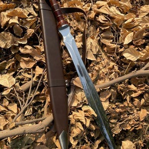 Könnte beinhalten: Ein zweischneidiges Gladius-Schwert mit einer polierten Stahlklinge, einem dunkelbraunen Holzgriff mit silberfarbenen Metallringen und einer passenden dunkelbraunen Lederscheide mit silberfarbener Metallspitze und integriertem Lederriemen.