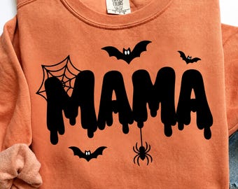 Pode incluir: Uma camisola de gola redonda laranja com uma impressão gráfica preta da palavra MAMA numa fonte a escorrer. O design inclui uma teia de aranha, dois morcegos com olhos brancos e uma aranha pendurada. Etiqueta da marca Comfort Colors.