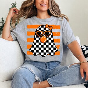 Pode incluir: Uma t-shirt cinzenta mesclada com um gr&aacute;fico de um fantasma com padr&atilde;o xadrez preto e branco. O fantasma usa &oacute;culos de sol laranja em forma de cora&ccedil;&atilde;o, tem uma bolha laranja e um pequeno la&ccedil;o laranja na cabe&ccedil;a, sobre riscas laranja.
