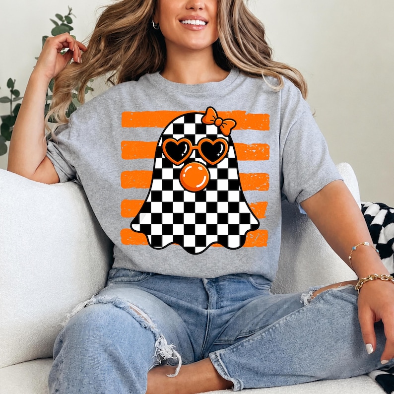 Pode incluir: Uma t-shirt cinzenta mesclada com um gr&aacute;fico de um fantasma com padr&atilde;o xadrez preto e branco. O fantasma usa &oacute;culos de sol laranja em forma de cora&ccedil;&atilde;o, tem uma bolha laranja e um pequeno la&ccedil;o laranja na cabe&ccedil;a, sobre riscas laranja.