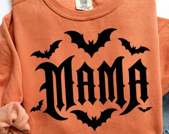 Pode incluir: Uma camisola laranja de manga comprida com uma impressão gráfica preta da palavra MAMA numa fonte de estilo gótico, rodeada por cinco silhuetas de morcegos pretos de vários tamanhos. A peça tem um decote redondo e uma etiqueta da marca Comfort Colors.