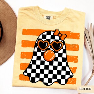 Pode incluir: Uma t-shirt de manga curta amarelo-p&aacute;lido com um gr&aacute;fico de um fantasma com um padr&atilde;o axadrezado preto e branco. O fantasma usa &oacute;culos de sol cor de laranja em forma de cora&ccedil;&atilde;o e um la&ccedil;o cor de laranja, com uma bolha cor de laranja sob os olhos sobre riscas cor de laranja.
