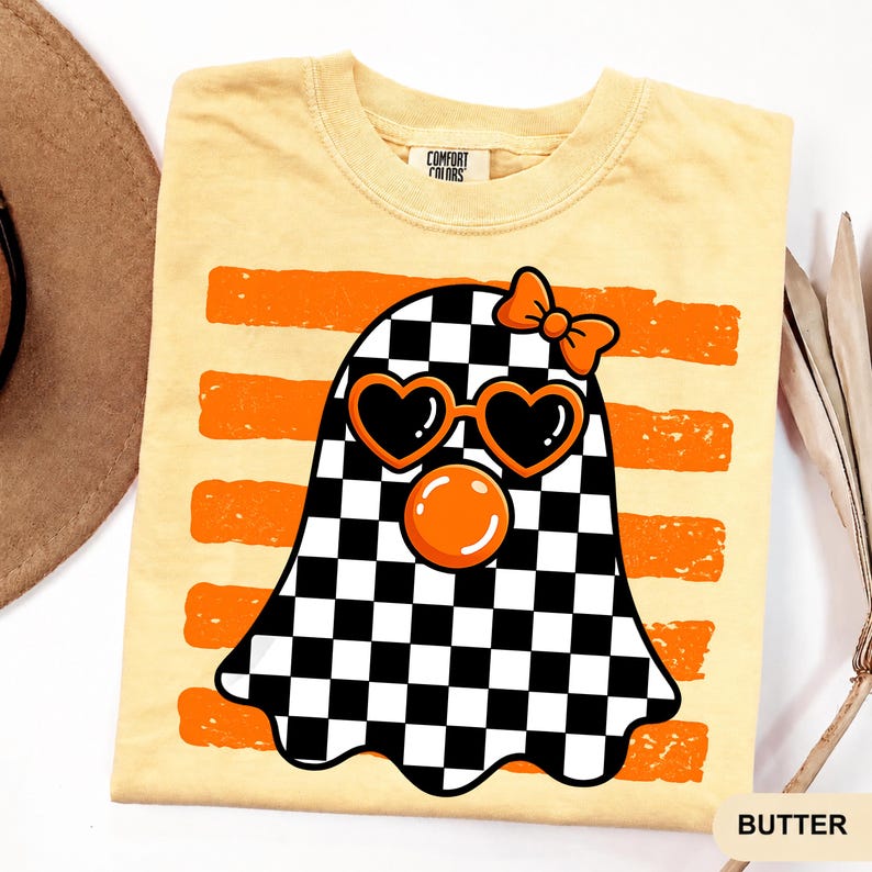 Pode incluir: Uma t-shirt de manga curta amarelo-p&aacute;lido com um gr&aacute;fico de um fantasma com um padr&atilde;o axadrezado preto e branco. O fantasma usa &oacute;culos de sol cor de laranja em forma de cora&ccedil;&atilde;o e um la&ccedil;o cor de laranja, com uma bolha cor de laranja sob os olhos sobre riscas cor de laranja.