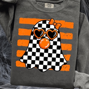 Pode incluir: Uma camisola de manga comprida cinzento-carv&atilde;o com o gr&aacute;fico de um fantasma com padr&atilde;o axadrezado preto e branco. O fantasma usa &oacute;culos de sol cor de laranja em forma de cora&ccedil;&atilde;o, um la&ccedil;o cor de laranja e tem uma bolha cor de laranja. Atr&aacute;s h&aacute; riscas.