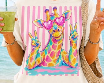 Pode incluir: Uma t-shirt de cor creme com um gráfico de uma girafa colorida a usar óculos de sol cor-de-rosa em forma de coração. A girafa faz sinais de paz com ambas as patas dianteiras sobre um fundo de riscas verticais cor-de-rosa. O design é vibrante.