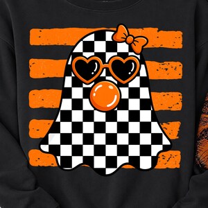 Pode incluir: Uma camisola preta com o gr&aacute;fico de um fantasma com padr&atilde;o xadrez preto e branco. O fantasma usa &oacute;culos de sol cor de laranja em forma de cora&ccedil;&atilde;o, um la&ccedil;o cor de laranja e est&aacute; a soprar uma bolha cor de laranja. O fundo tem riscas horizontais.