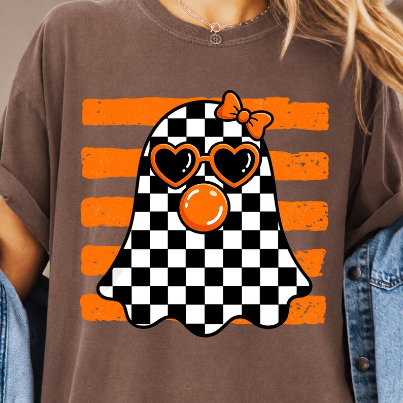 Pode incluir: Uma t-shirt castanha com um gr&aacute;fico de um fantasma com um padr&atilde;o axadrezado preto e branco. O fantasma usa &oacute;culos de sol cor de laranja em forma de cora&ccedil;&atilde;o e um la&ccedil;o cor de laranja, e est&aacute; a soprar uma bolha cor de laranja. O fundo tem riscas.