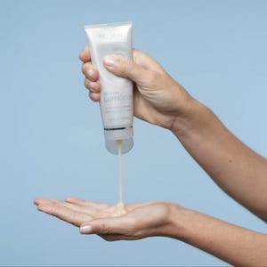 May include: A white squeeze tube of Nu Skin LumiSpa activating cleanser with silver accents. The tube features circular graphic branding and text, dispensing a thick, light-colored gel product from the bottom opening into an open palm.
