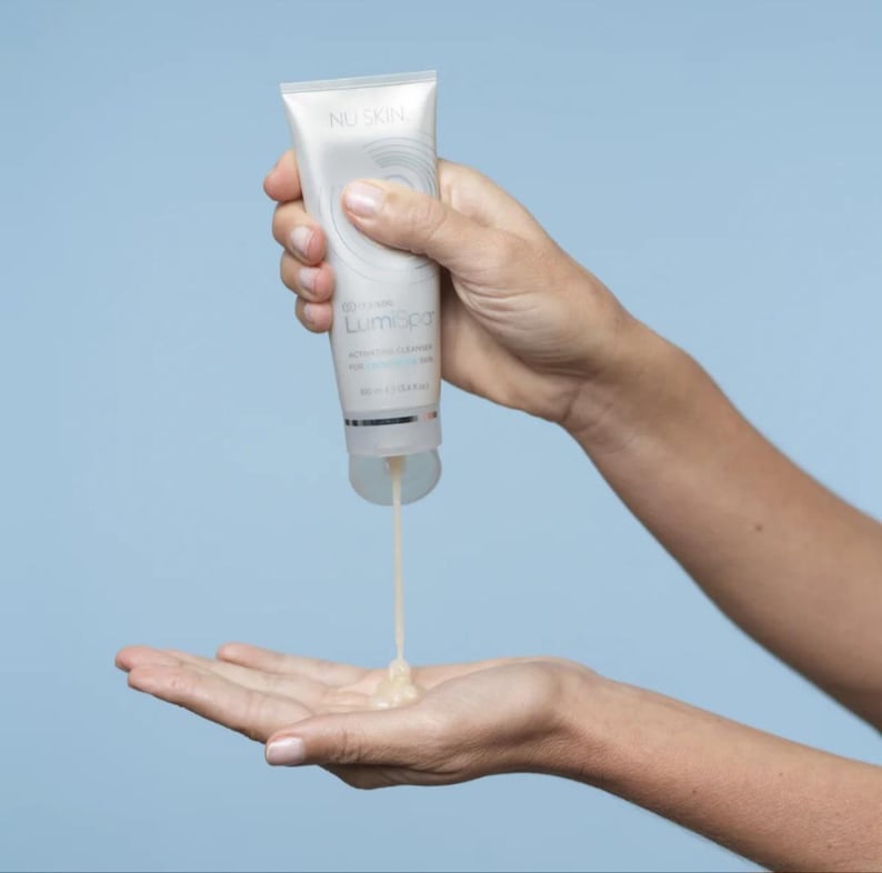 May include: A white squeeze tube of Nu Skin LumiSpa activating cleanser with silver accents. The tube features circular graphic branding and text, dispensing a thick, light-colored gel product from the bottom opening into an open palm.
