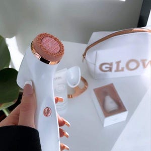 Peut inclure: Une brosse nettoyante pour le visage blanche et or rose avec une t&ecirc;te en silicone rose textur&eacute;e. &Agrave; proximit&eacute; se trouvent une trousse de toilette blanche avec le mot GLOW en lettres or rose, un petit tube blanc, un support et un embout en bo&icirc;te.