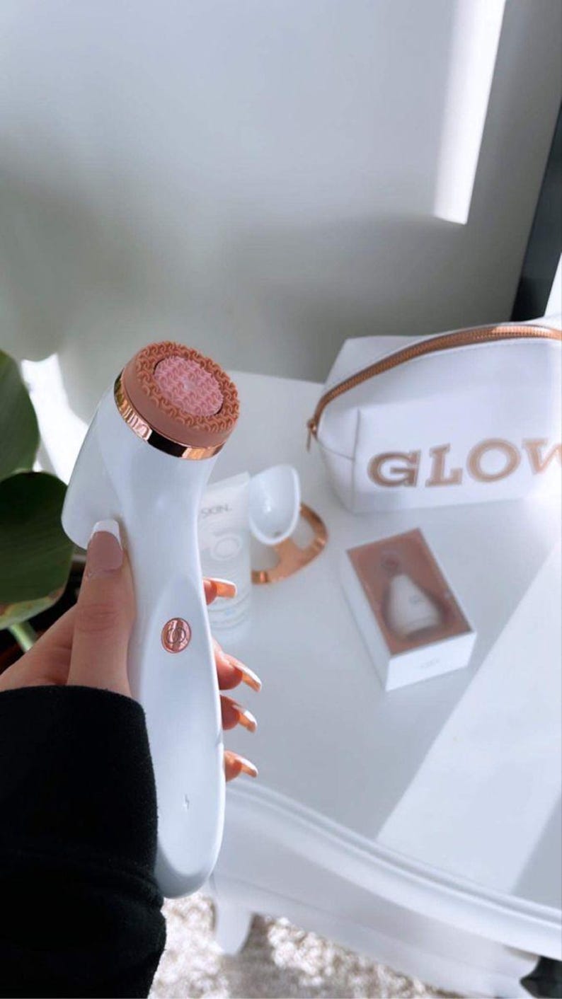 Peut inclure: Une brosse nettoyante pour le visage blanche et or rose avec une t&ecirc;te en silicone rose textur&eacute;e. &Agrave; proximit&eacute; se trouvent une trousse de toilette blanche avec le mot GLOW en lettres or rose, un petit tube blanc, un support et un embout en bo&icirc;te.