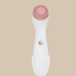 Peut inclure: Un appareil de nettoyage facial blanc avec une base circulaire en or rose et un anneau assorti autour de la t&ecirc;te de brosse. L'appareil pr&eacute;sente une surface de brosse en silicone rose textur&eacute;e, un bouton d'alimentation central et des voyants.