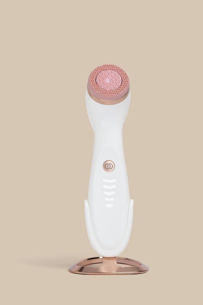 Peut inclure: Un appareil de nettoyage facial blanc avec une base circulaire en or rose et un anneau assorti autour de la t&ecirc;te de brosse. L'appareil pr&eacute;sente une surface de brosse en silicone rose textur&eacute;e, un bouton d'alimentation central et des voyants.