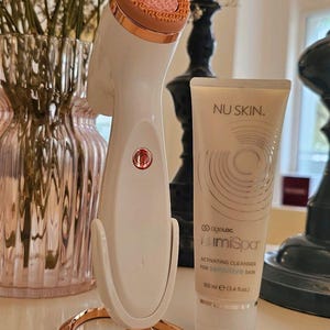 Peut inclure: Un appareil de nettoyage facial blanc et or rose avec une t&ecirc;te en silicone rose textur&eacute;e sur un support assorti, accompagn&eacute; d'un tube de 100 ml de nettoyant activateur Nu Skin ageLOC LumiSpa pour peau sensible avec un motif graphique circulaire.