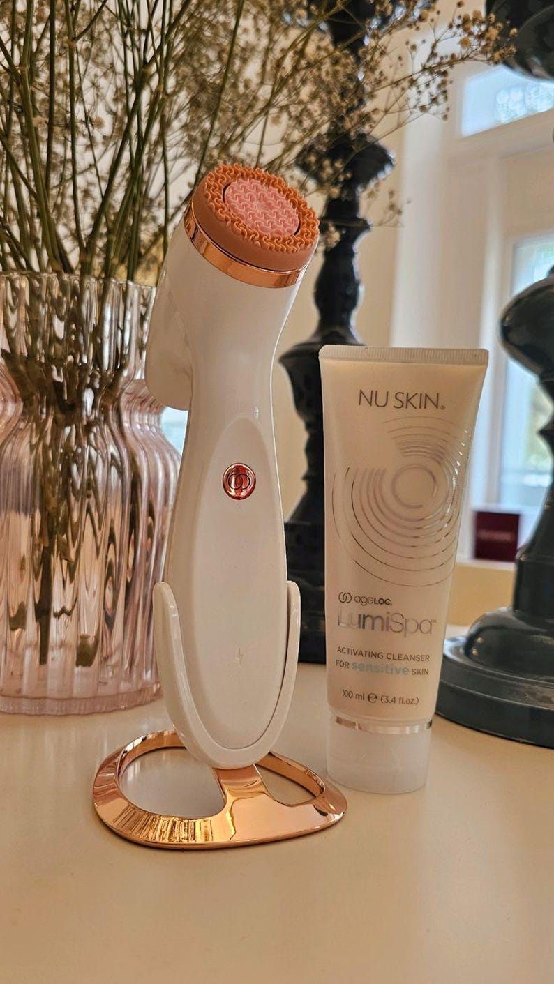 Peut inclure: Un appareil de nettoyage facial blanc et or rose avec une t&ecirc;te en silicone rose textur&eacute;e sur un support assorti, accompagn&eacute; d'un tube de 100 ml de nettoyant activateur Nu Skin ageLOC LumiSpa pour peau sensible avec un motif graphique circulaire.