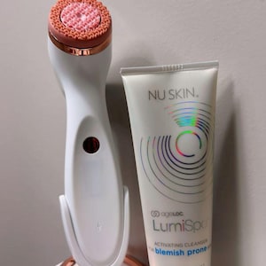 Peut inclure: Un appareil de nettoyage facial blanc et or rose sur son socle assorti, accompagn&eacute; d'un tube de 100 ml de nettoyant activateur Nu Skin ageLOC LumiSpa pour peau &agrave; imperfections. Le tube pr&eacute;sente un motif circulaire holographique et du texte noir.
