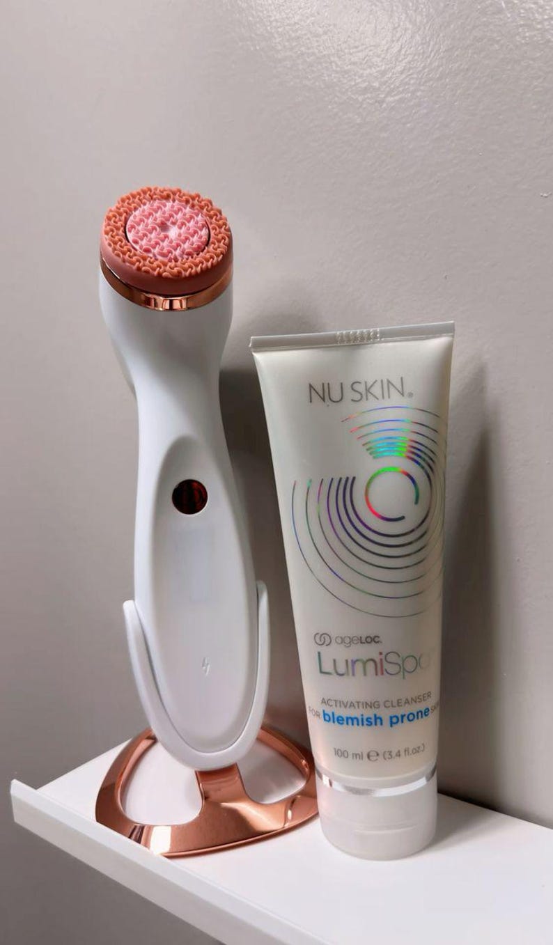 Peut inclure: Un appareil de nettoyage facial blanc et or rose sur son socle assorti, accompagn&eacute; d'un tube de 100 ml de nettoyant activateur Nu Skin ageLOC LumiSpa pour peau &agrave; imperfections. Le tube pr&eacute;sente un motif circulaire holographique et du texte noir.