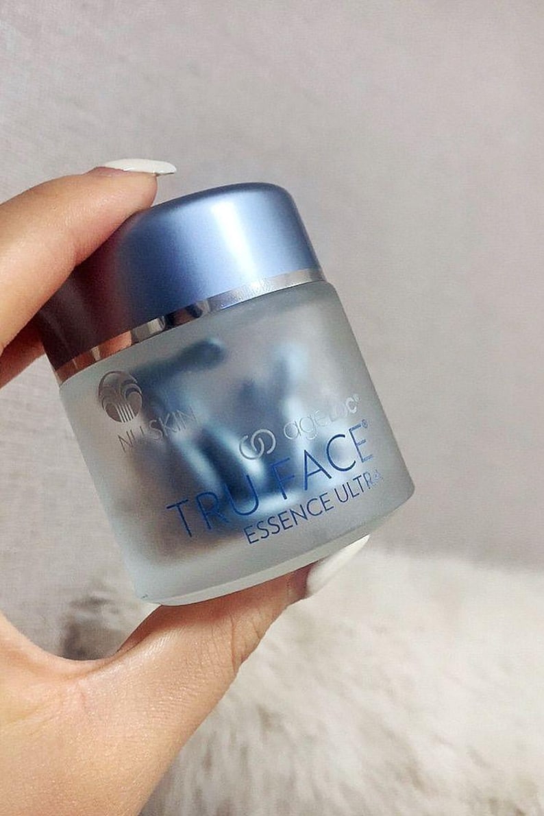 Pu&ograve; includere: Un barattolo in vetro smerigliato con coperchio blu metallizzato contenente capsule blu. L'etichetta riporta il logo Nu Skin e la scritta ageLOC TRU FACE ESSENCE ULTRA in blu. Il contenitore ha una forma cilindrica e una finitura semitrasparente.