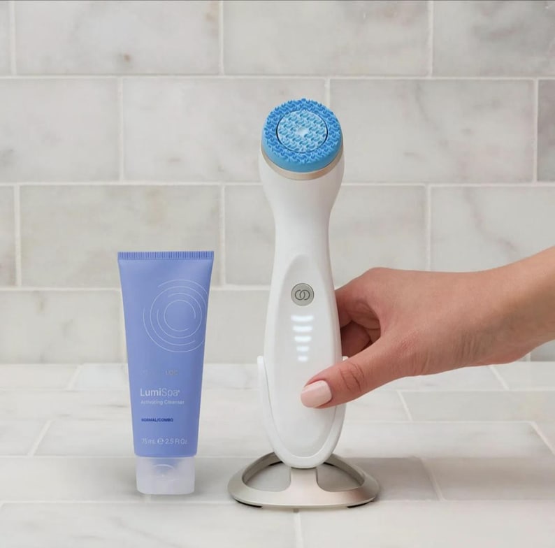 May include: A white facial cleansing device with a blue silicone head on a silver stand, accompanied by a light purple tube of LumiSpa Activating Cleanser labeled 75 ml and 2.5 fl oz, featuring a circular graphic design on the front.