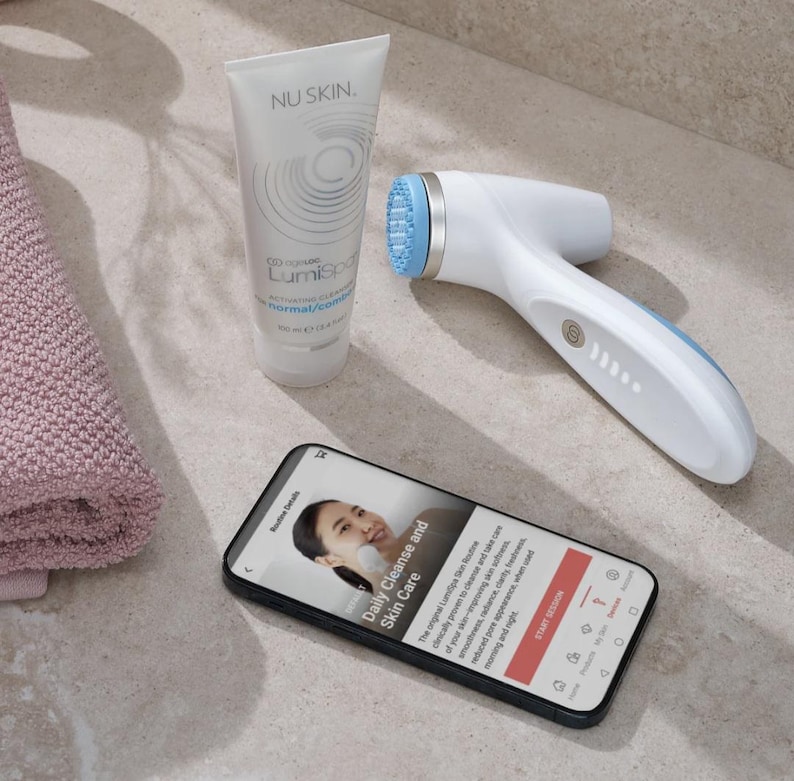 May include: A white Nu Skin LumiSpa facial cleansing device with a light blue silicone head, a tube of Nu Skin activating cleanser, and a smartphone displaying a skin care routine app interface with text about daily cleanse and skin care.