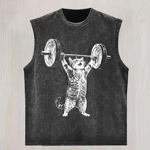 May include: A black sleeveless shirt with a distressed, washed-out texture. The center features a white graphic illustration of a tabby cat standing on its hind legs while lifting a heavy barbell with weights above its head with both paws.