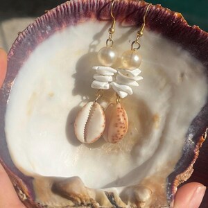 May include: A pair of dangle earrings featuring gold-tone hooks, a single cream-colored pearl bead, a stack of three white shell discs, and a single tan and brown spotted cowrie shell at the base, all suspended from a gold-tone metal wire.