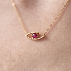 May include: A gold necklace featuring an eye-shaped pendant with a central round-cut ruby gemstone. The pendant is suspended from a delicate gold chain, creating a simple and refined piece of jewelry that rests against the skin.