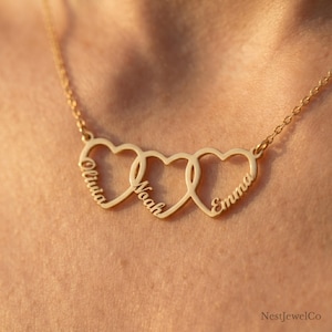 May include: A gold-toned necklace featuring three interlocking heart-shaped pendants. Each heart is engraved with a name in a cursive script: Olivia on the left, Noah in the center, and Emma on the right. The piece includes a delicate chain.