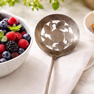 May include: A silver serving spoon with a decorative floral design next to a white bowl filled with fresh berries. The bowl contains raspberries, blueberries, and blackberries, with mint leaves for garnish. The spoon rests on a white cloth.
