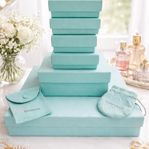May include: A stack of light blue gift boxes of varying sizes, with a matching pouch and drawstring bag. The boxes are arranged on a white surface, with a bouquet of white roses and perfume bottles in the background. The Tiffany & Co. logo is visible.