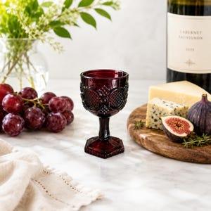 May include: A deep red pressed glass goblet featuring a textured geometric pattern on the bowl and a hexagonal base. The glass has a short stem and a thick, ornate rim, showcasing a vintage-inspired design with intricate embossed detailing.