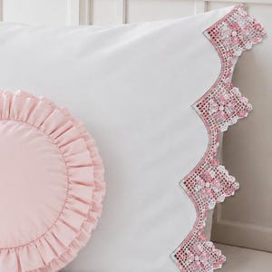 May include: A white rectangular pillowcase featuring a scalloped edge trimmed with intricate white and pink lace, paired with a round, light pink decorative pillow that has a gathered, ruffled border around its entire circular perimeter.