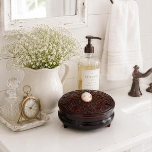 May include: A dark brown, round, lidded jewelry box with an oval cameo accent on top, placed next to a white ceramic pitcher filled with baby's breath, a small gold-toned clock, a glass perfume bottle, and a bottle of lavender hand soap.