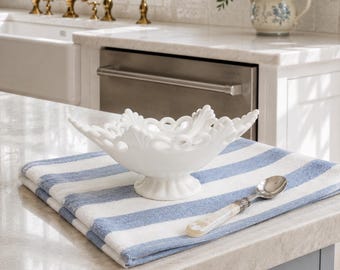May include: A white milk glass bowl with a scalloped, openwork rim and a pedestal base, resting on a folded white and blue striped cotton towel next to a silver-toned spoon with a pearlescent handle, all displayed on a kitchen worktop.