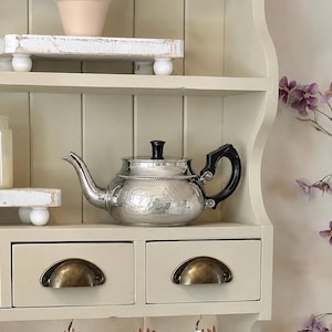 May include: A silver teapot with a black handle and lid sits on a shelf. The teapot has a hammered texture and a decorative chain around the lid. The shelf is part of a cream-colored cabinet with drawers and brass-colored handles.