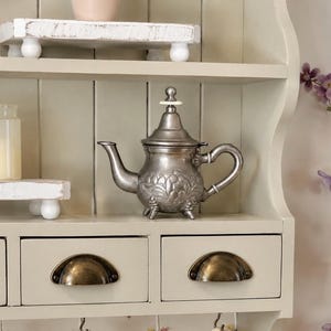 May include: A silver teapot with floral detailing sits on a shelf. The shelf is part of a cream-colored cabinet with drawers. The cabinet has a vintage aesthetic, with a small jar and decorative wooden risers.