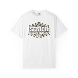 May include: A white short-sleeve t-shirt featuring a central hexagonal graphic with a camouflage pattern. The text SENIOR CLASS OF 2027 is printed in a bold, white, block-style font across the center of the camouflage hexagon design.