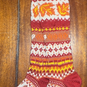 May include: A pair of knee-high knitted socks featuring a multi-colored fair isle pattern in shades of red, orange, yellow, and cream. The design includes repeating motifs of autumn leaves and acorns, with a ribbed cuff and a solid red toe and heel.