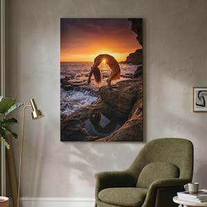 May include: A rectangular wall art print featuring a person in a yellow dress performing a backbend on a rocky coastal cliff at sunset. The sun is positioned directly behind the person, casting a warm orange glow over the ocean and rocks.