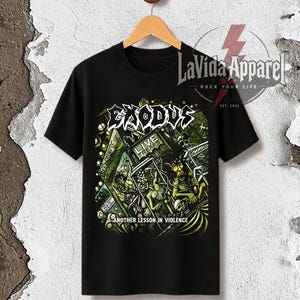 Può includere: Una t-shirt nera a maniche corte con una stampa grafica che riporta il testo Exodus e Another Lesson in Violence. Il design include illustrazioni verdi e gialle di figure all'interno di una struttura a gabbia con il logo LaVida Apparel.